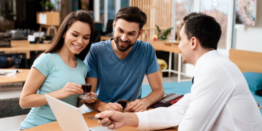 Vendedor aponta para a tela do notebook enquanto dois clientes olham atentamente e sorriem com xícaras de café nas mãos.