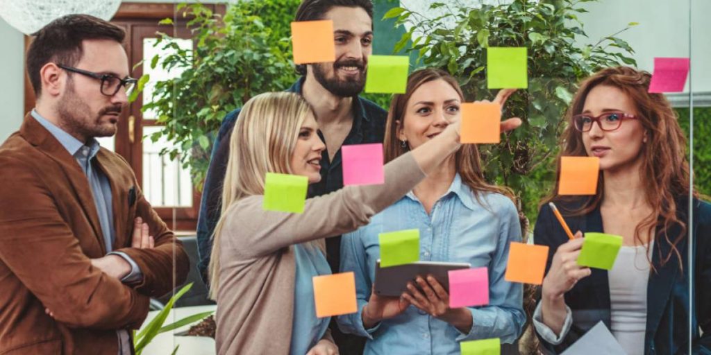 grupo de colaboradores reunidos de pé discutem as perguntas da pesquisa de clima organizacional em uma parede de vidro com post-its.