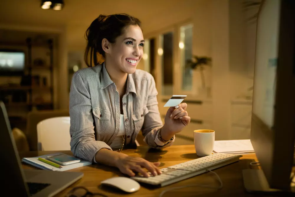 mulher sorrindo segurando um cartão de crédito e mexendo no computador para falar sobre programa de pontos