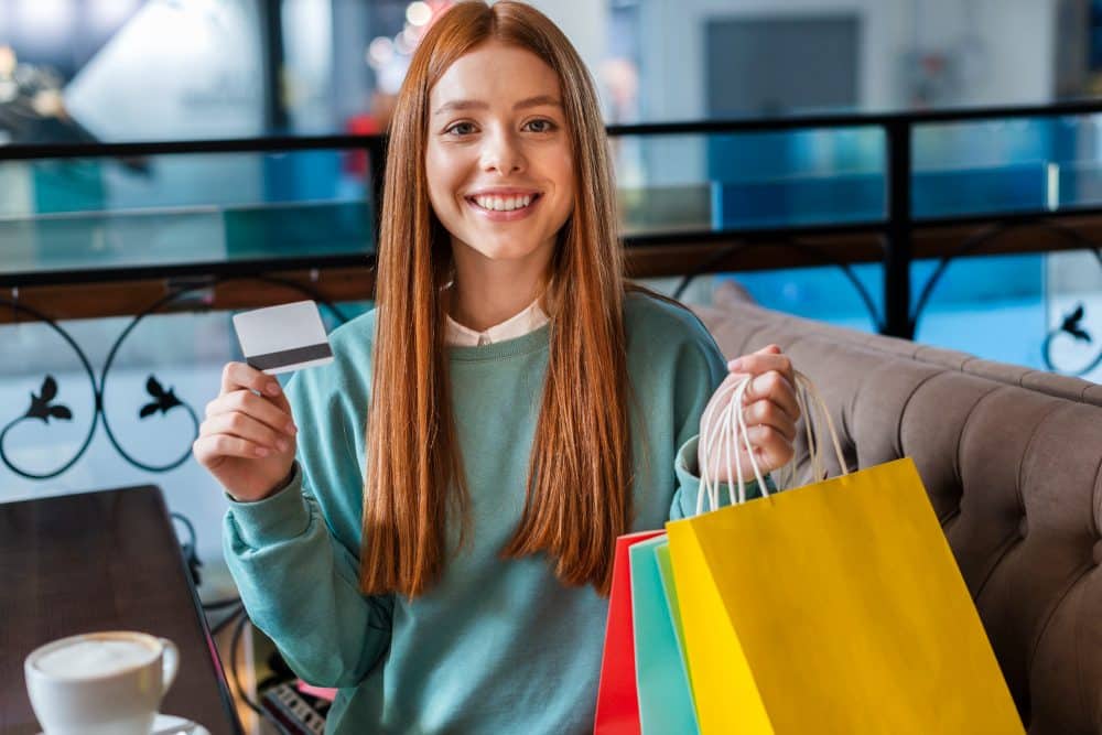 Mulher jovem sorrindo enquanto segura sacolas de compras em uma mão e cartão de benefícios em outra.
