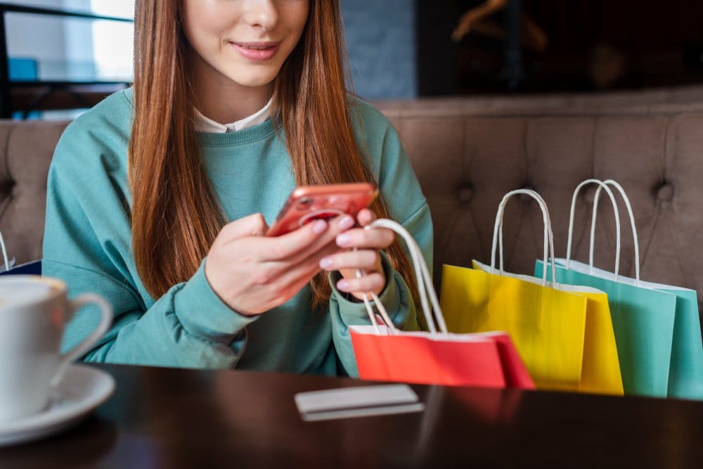 Mulher sentada ao lado de sacolas de compras enquanto faz compras online pelo celular.