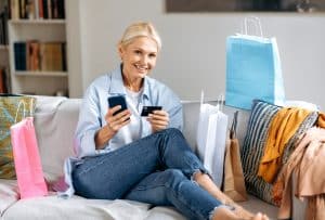 Mulher sentada em um sofá com cartão de crédito e celular em mão. No sofá contém várias sacolas de compras