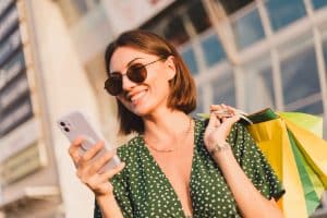 Uma mulher sorridente, de cabelo curto e óculos de sol redondos, olha para seu smartphone enquanto segura sacolas de compras no ombro