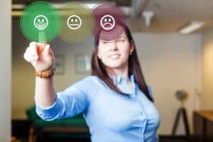 Uma mulher com uma camisa azul clara e cabelos escuros, com o foco em seu braço direito estendido e mão apontando para frente. Na ponta de seu dedo indicador, há três círculos virtuais translúcidos com ícones de emoji de feedback