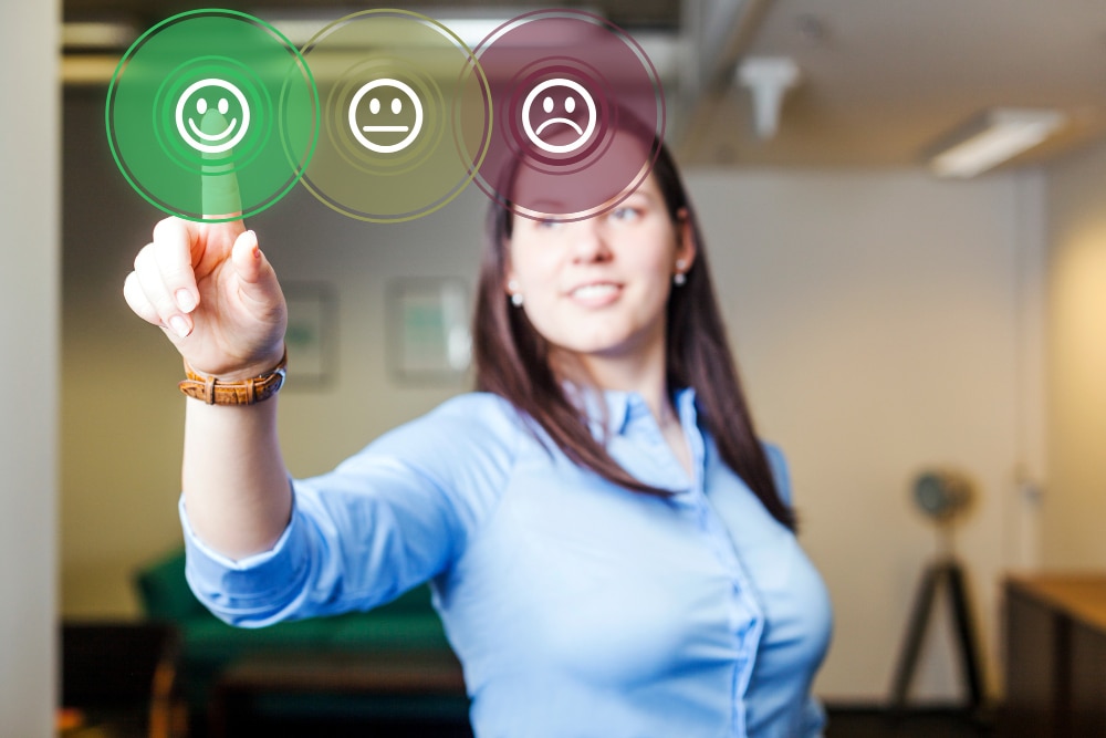 Uma mulher com uma camisa azul clara e cabelos escuros, com o foco em seu braço direito estendido e mão apontando para frente. Na ponta de seu dedo indicador, há três círculos virtuais translúcidos com ícones de emoji de feedback