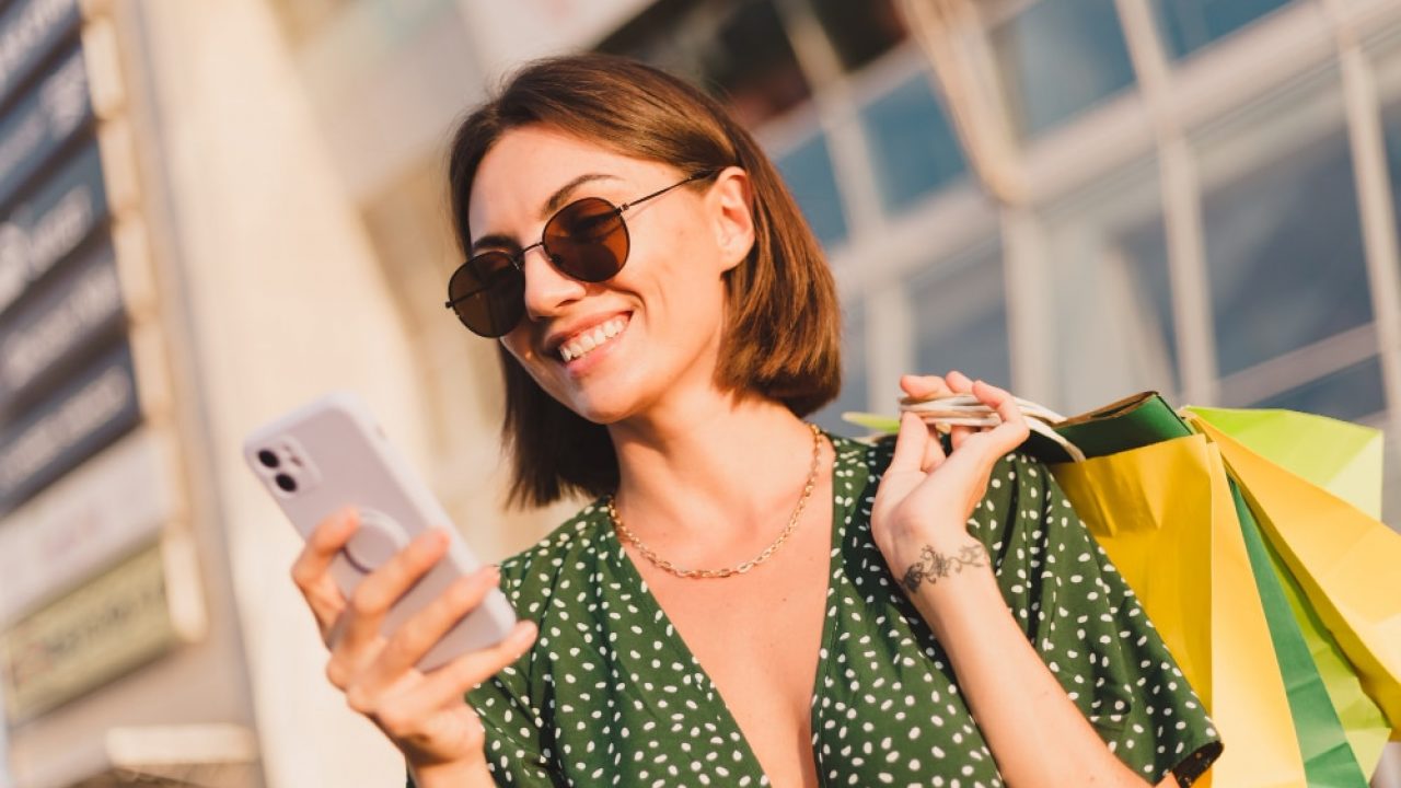 Uma mulher sorridente, de cabelo curto e óculos de sol redondos, olha para seu smartphone enquanto segura sacolas de compras no ombro Uma mulher sorridente, de cabelo curto e óculos de sol redondos, olha para seu smartphone enquanto segura sacolas de compras no ombro