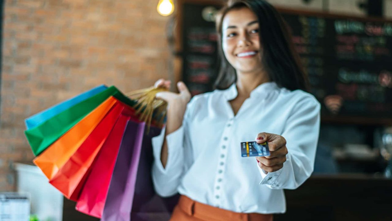 mulher-com-blusa-branca-com-um-cartao-em-uma-das-mao-e-na-outra-sacolas-de-compras.