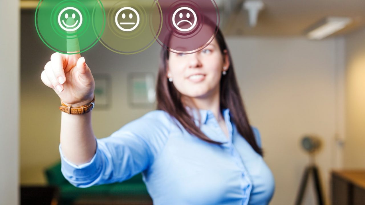 Uma mulher com uma camisa azul clara e cabelos escuros, com o foco em seu braço direito estendido e mão apontando para frente. Na ponta de seu dedo indicador, há três círculos virtuais translúcidos com ícones de emoji de feedback